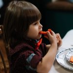 Criança pequena come cenoura e couve-flor à mesa, num ambiente interior.