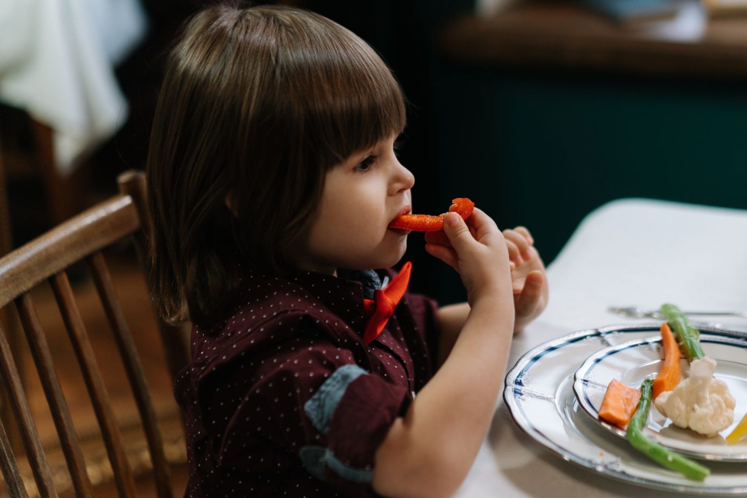 Criança pequena come cenoura e couve-flor à mesa, num ambiente interior.
