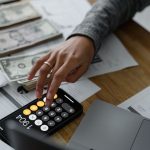 Pessoa usa a calculadora do celular entre dinheiro e documentos financeiros sobre uma mesa.