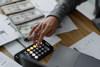 Pessoa usa a calculadora do celular entre dinheiro e documentos financeiros sobre uma mesa.