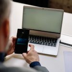 Profissional usa o celular para realizar um pagamento digital ao lado de um laptop em um escritório