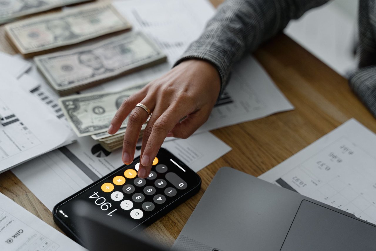 Pessoa usa a calculadora do celular entre dinheiro e documentos financeiros sobre uma mesa.